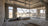 An unfinished concrete room with exposed ceiling pipes, large windows, a metal support frame, and a small white bench or platform near the center. The space is shown in a real architectural environment with desert scenery visible outside.